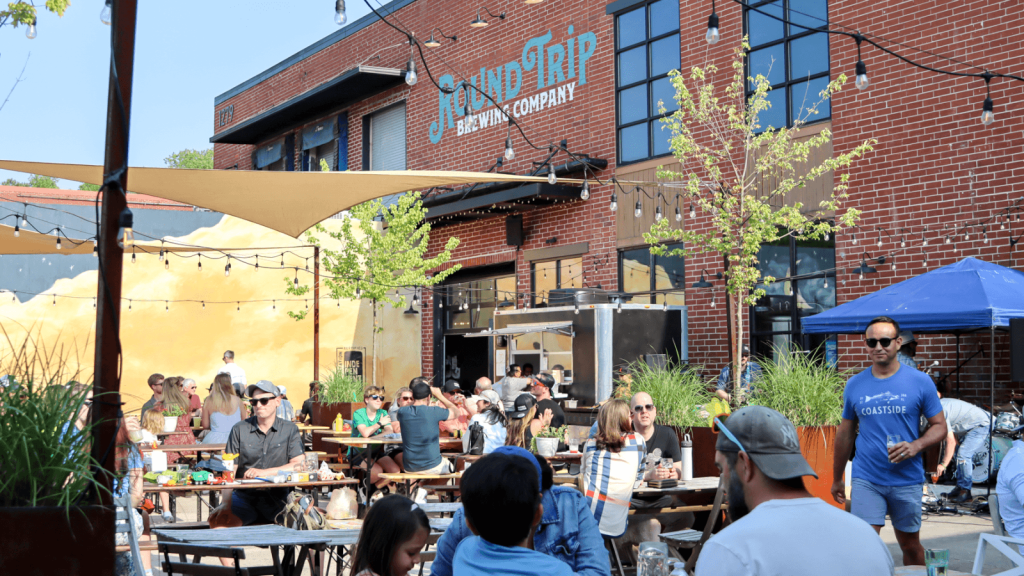 A bustling outdoor patio at Round Trip Brewing Company, filled with people enjoying food, drinks, and live music under colorful umbrellas and string lights in Atlanta, Georgia.