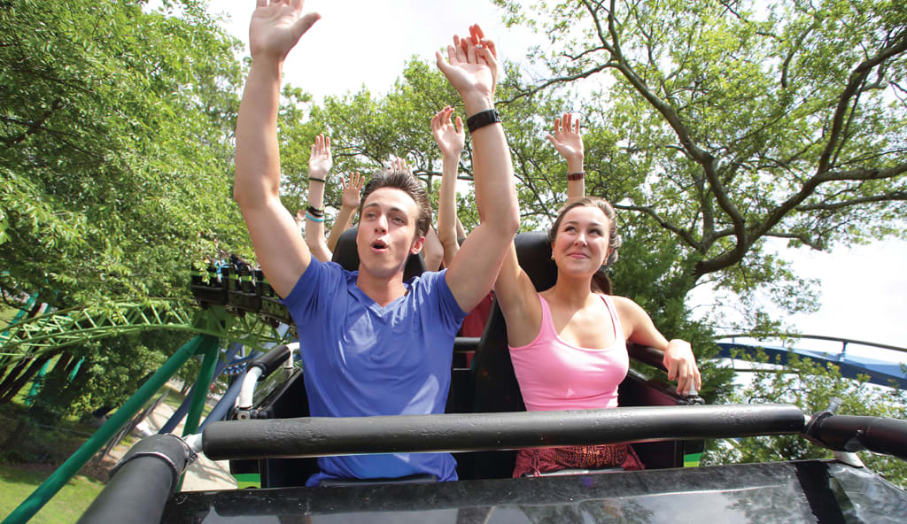 A couple rides a roller coaster, arms raised in excitement, surrounded by lush green trees in Atlanta, Georgia.