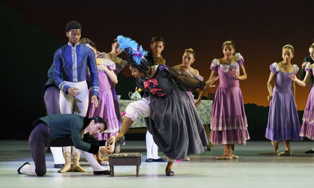 A ballet scene featuring a man in a blue coat kneeling to adjust a ballerina's slipper, surrounded by dancers in colorful period costumes in Atlanta, Georgia.