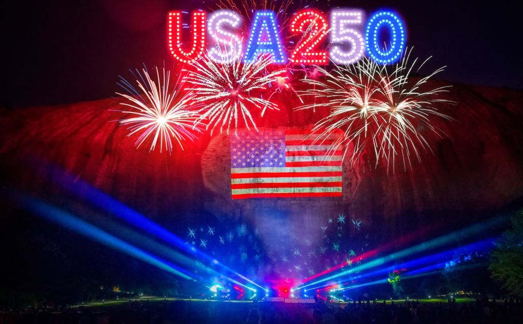 A vibrant fireworks display celebrating USA250 with colorful bursts and a glowing American flag projected on a rocky cliff face in Atlanta, Georgia.