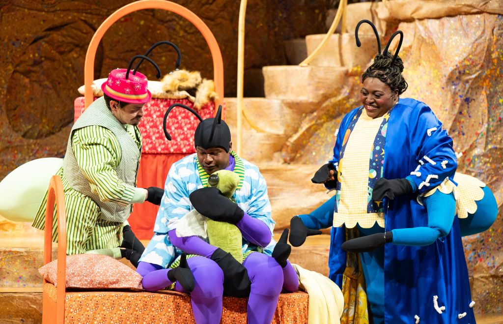 Three performers in whimsical costumes with insect-like antennae engage in a playful scene on a stage set resembling a giant bed in Atlanta, Georgia.