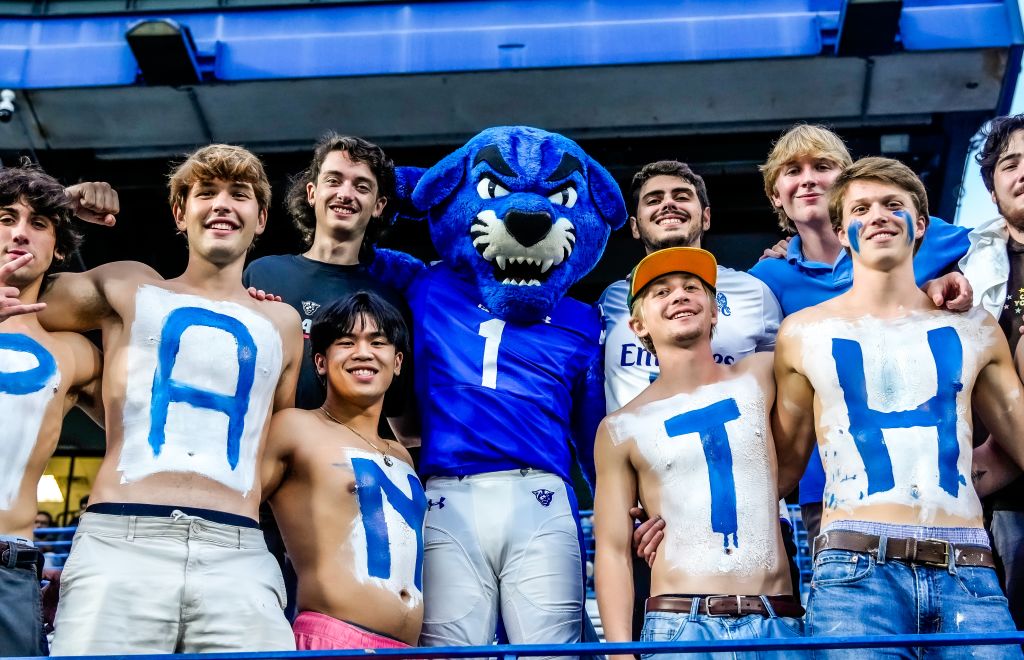 A group of shirtless young men with painted bodies and a blue mascot celebrate at a sports event in Atlanta, Georgia.