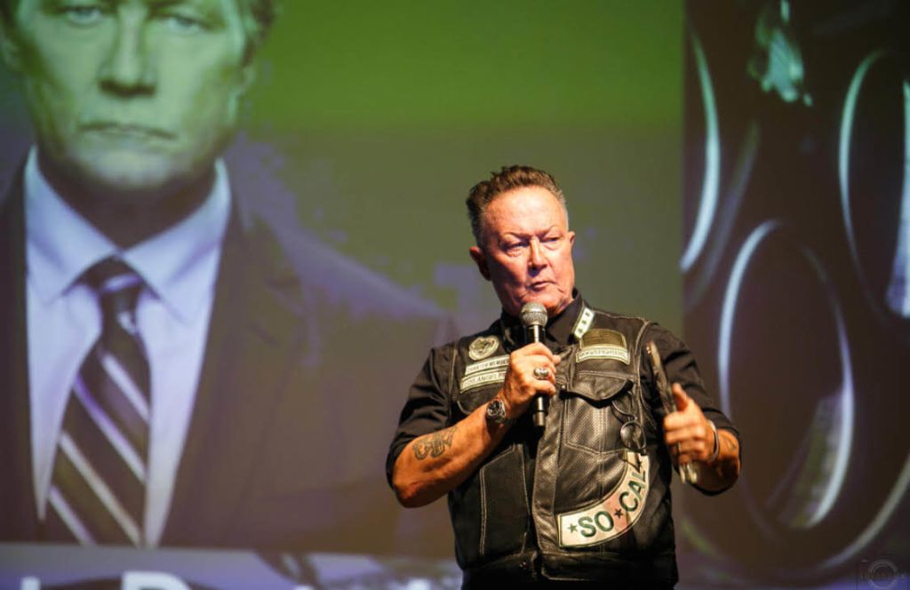 A man in a leather vest with patches is speaking into a microphone on stage, with a projected image of a man in a suit behind him in Atlanta, Georgia.