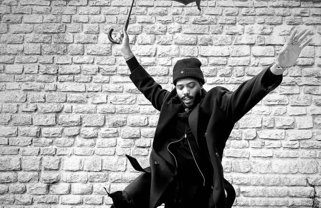 A man in a black coat and beanie dances with an umbrella against a brick wall in Atlanta, Georgia.