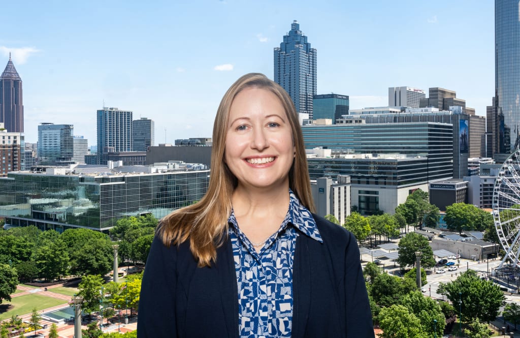 A smiling woman stands in front of a city skyline with skyscrapers and a Ferris wheel in Atlanta, Georgia.
