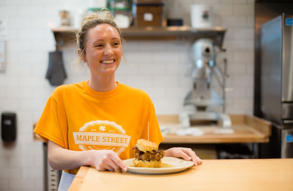 A smiling woman in an orange Maple Street t-shirt holds a plate with a sandwich in Atlanta, Georgia.
