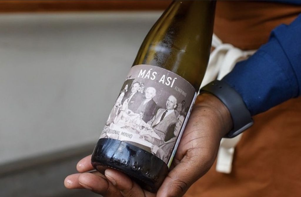 A hand holds a bottle of Más Asi wine with a label featuring a black and white photo of people at a table in Atlanta, Georgia.