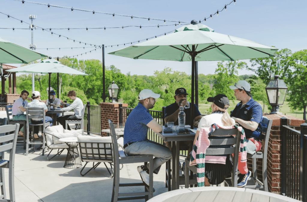 A group of people are enjoying a sunny day on an outdoor patio, seated at tables with green umbrellas, surrounded by lush greenery and string lights in Atlanta, Georgia.