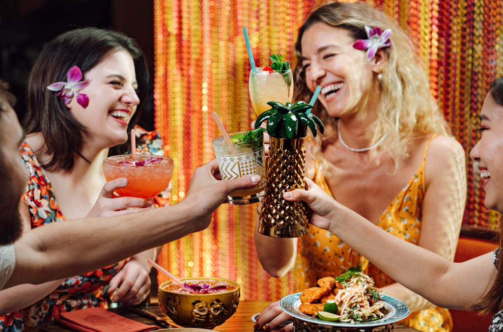 Four women in summer dresses laugh and clink tropical-themed cocktail glasses at a restaurant table in Atlanta, Georgia.