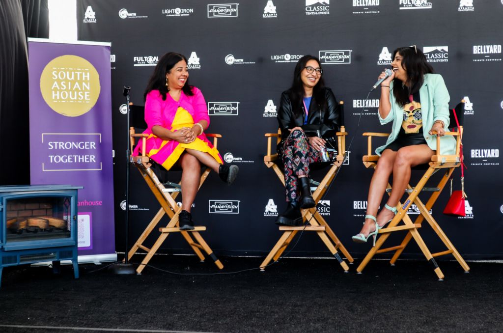 Three women sit on director's chairs on a stage, laughing and engaging in conversation at a South Asian House event in Atlanta, Georgia.
