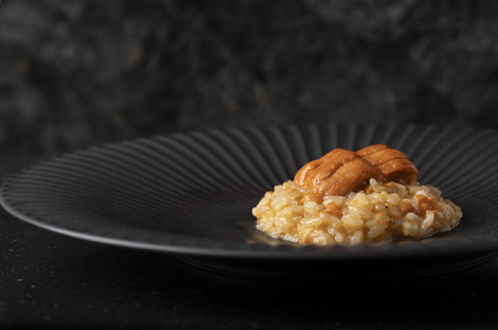 A small mound of creamy risotto topped with three pieces of bright orange uni (sea urchin) on a black plate in Atlanta, Georgia.