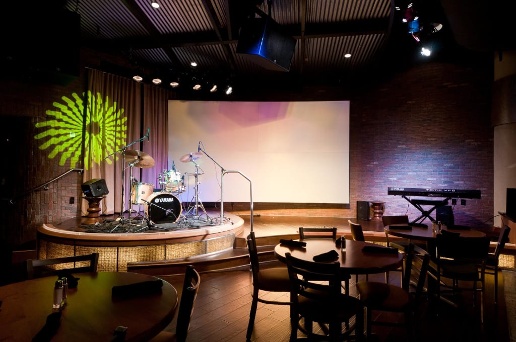 A dimly lit room with a stage featuring a drum set and keyboard, surrounded by tables and chairs in Atlanta, Georgia.