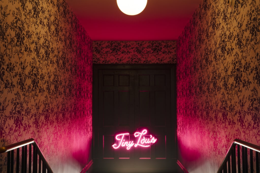 A dimly lit hallway with floral wallpaper leads to a door illuminated by a pink neon sign in Atlanta, Georgia.