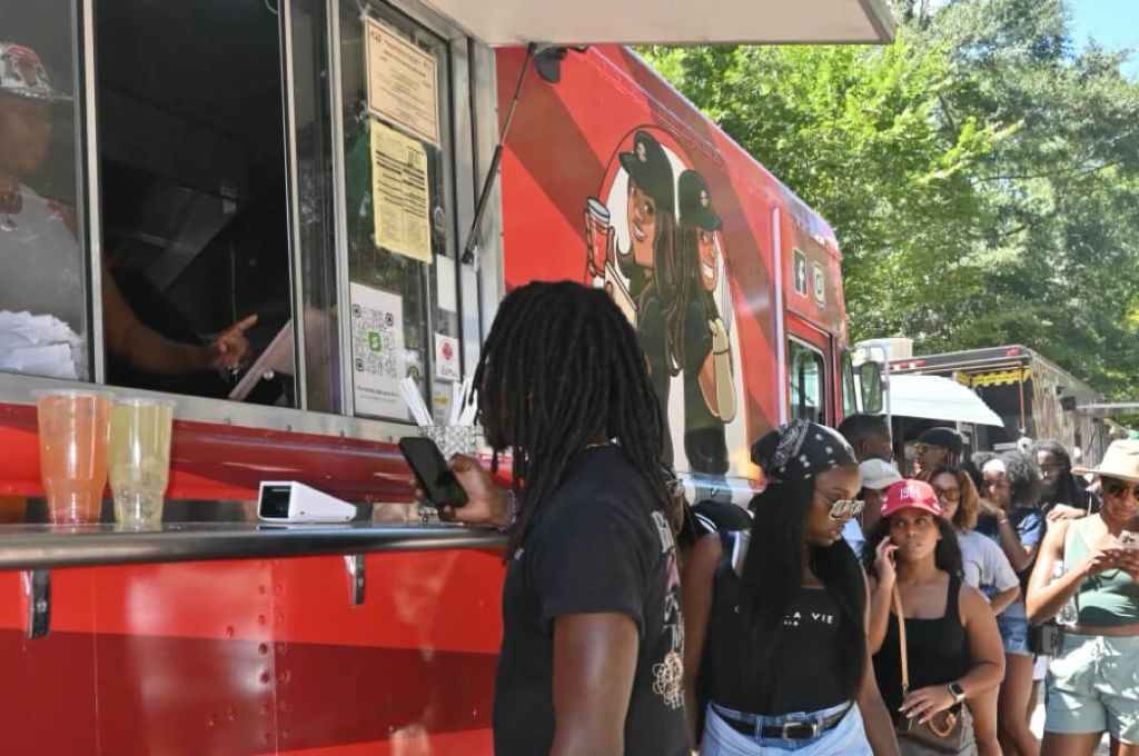 A vibrant red food truck with cartoon characters serves a diverse crowd of people enjoying a sunny day outdoors in Atlanta, Georgia.