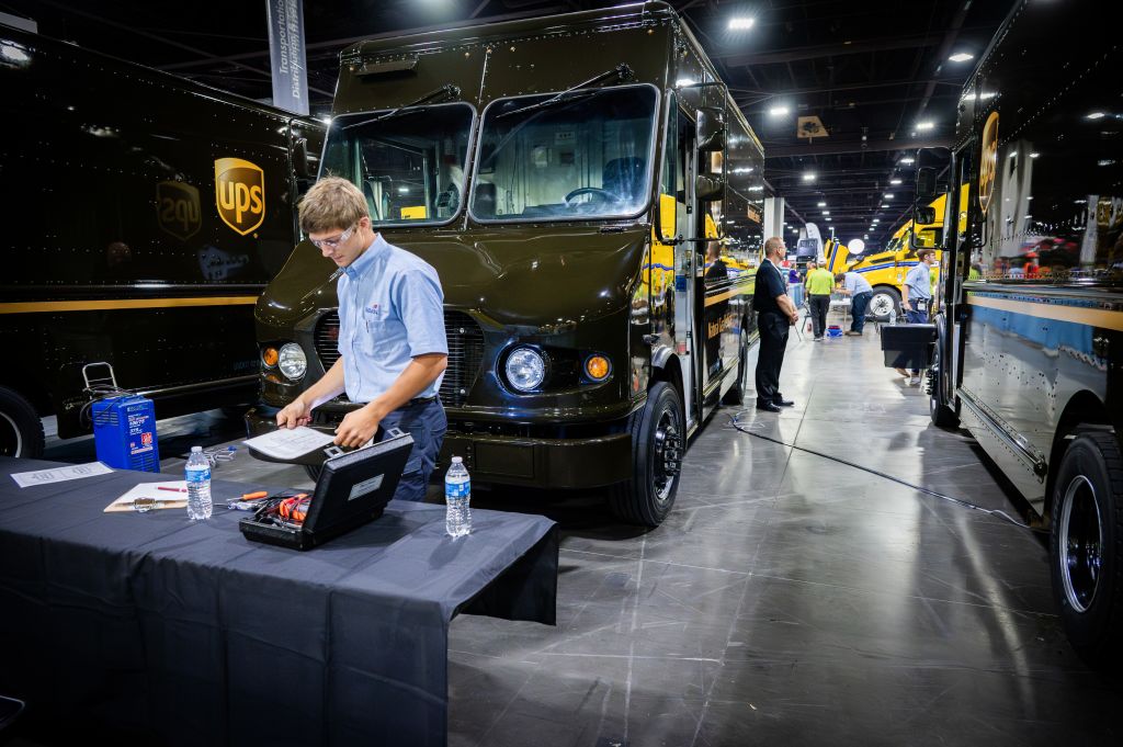 A UPS delivery truck is parked in a large exhibition hall, with a young man in a blue shirt inspecting documents at a table nearby in Atlanta, Georgia.