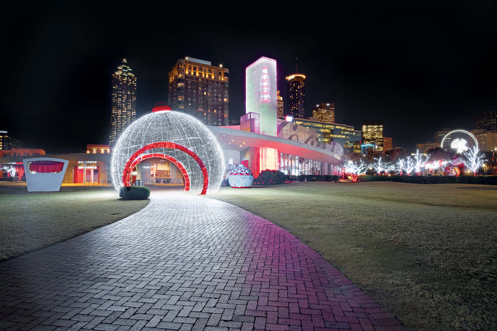 A nighttime scene featuring a large, illuminated dome with a red archway entrance, surrounded by a brick pathway and a city skyline in the background in Atlanta, Georgia.