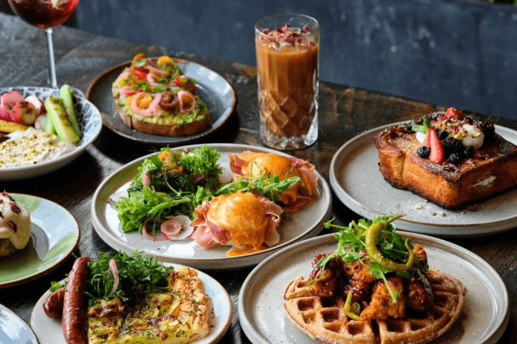 A variety of colorful and appetizing dishes, including waffles, eggs benedict, avocado toast, and a refreshing iced coffee, are artfully arranged on a rustic wooden table in Atlanta, Georgia.