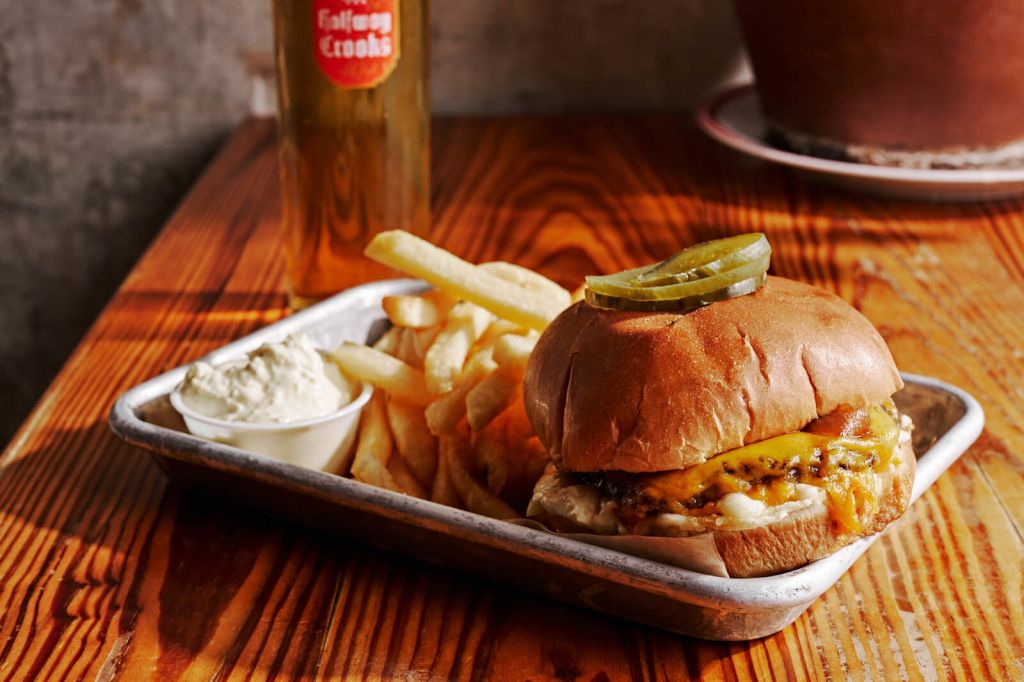 A mouthwatering cheeseburger with a pickle slice on top, served on a metal tray alongside crispy french fries, a small container of creamy sauce, and a tall glass of beer in Atlanta, Georgia.