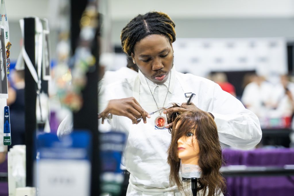 A young man with dreadlocks is carefully cutting hair on a mannequin at a cosmetology competition in Atlanta, Georgia.