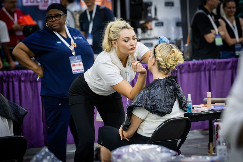 A makeup artist applies eyeliner to a client's eye at a beauty event, while another woman observes nearby in Atlanta, Georgia.