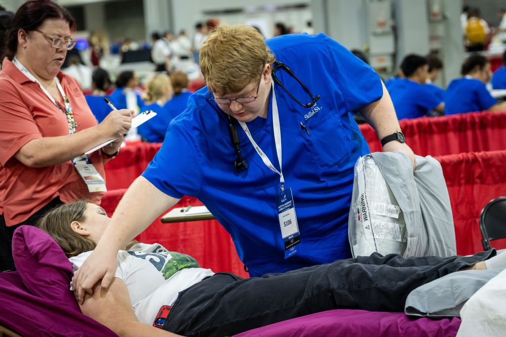 A medical professional in blue scrubs assists a patient lying on a bed, while another person in a red shirt takes notes nearby in Atlanta, Georgia.