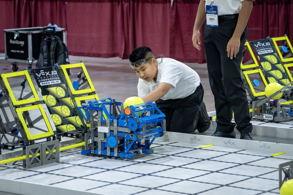 A young participant in a robotics competition carefully adjusts a blue and yellow robot designed to maneuver yellow balls on a grid-like playing field in Atlanta, Georgia.