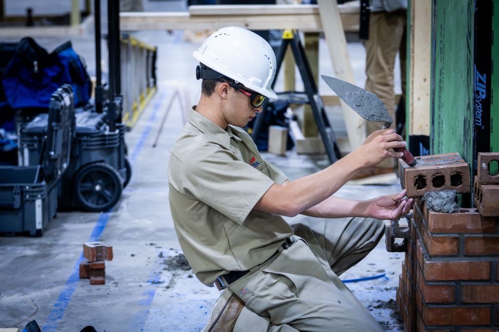 A construction worker in a white hard hat and safety glasses is kneeling to apply mortar to a brick wall with a trowel in Atlanta, Georgia.