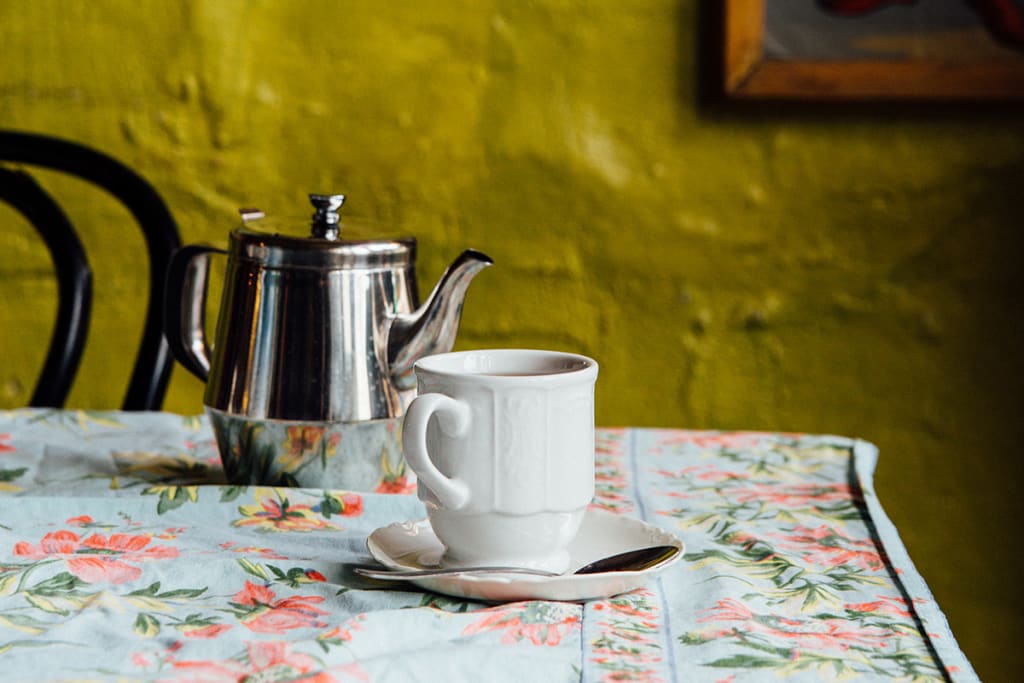 A white teacup and saucer with a silver spoon sit on a floral tablecloth next to a stainless steel teapot in Atlanta, Georgia.
