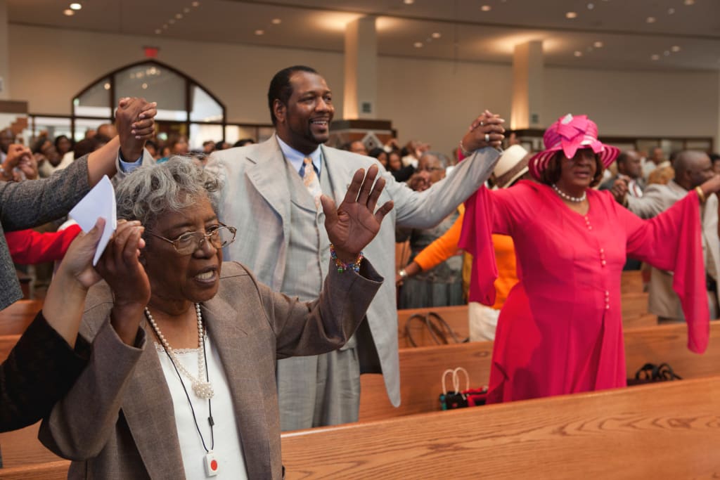 A diverse congregation of people, including an elderly woman in a brown jacket, a man in a suit, and a woman in a pink dress, raise their hands in unison during a church service in Atlanta, Georgia.