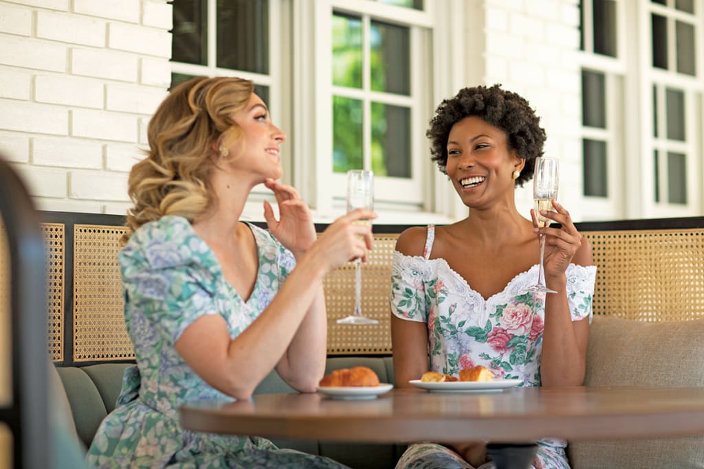 Ultimate Guide to Hotel Dining ATL Style 15 Two women in floral dresses enjoy a celebratory toast with champagne in a bright, airy setting in Atlanta, Georgia.