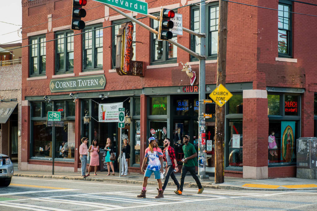 A bustling city street corner with pedestrians crossing, traffic lights, and a brick building with a tavern sign in Atlanta, Georgia.