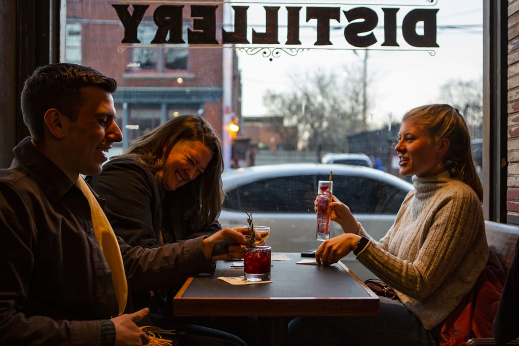 Three friends laugh and enjoy drinks together at a cozy bar table in Atlanta, Georgia.
