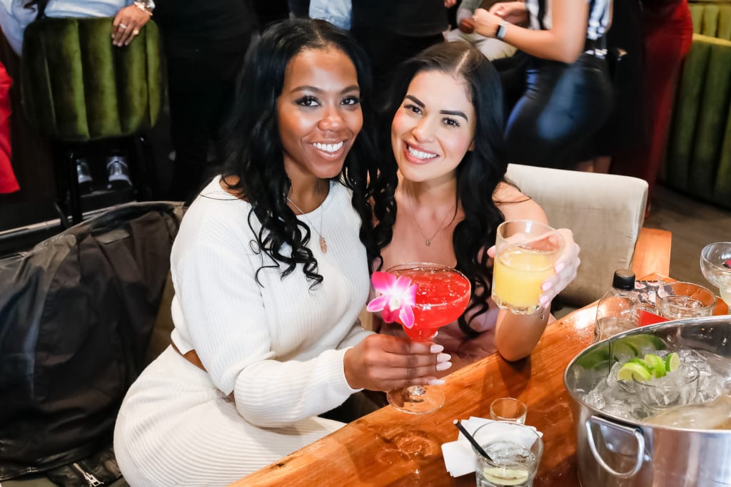 Two women enjoying colorful cocktails at a bar, smiling and posing for the camera in Atlanta, Georgia.
