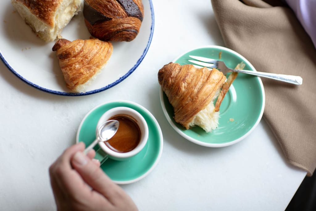A hand holds a spoon over a cup of espresso, with croissants and a fork on a green plate nearby in Atlanta, Georgia.