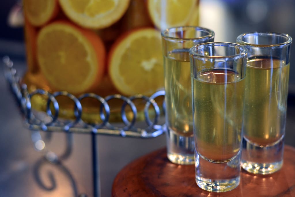 Three shot glasses filled with a golden liquid sit on a wooden surface next to a pitcher of orange slices in Atlanta, Georgia.