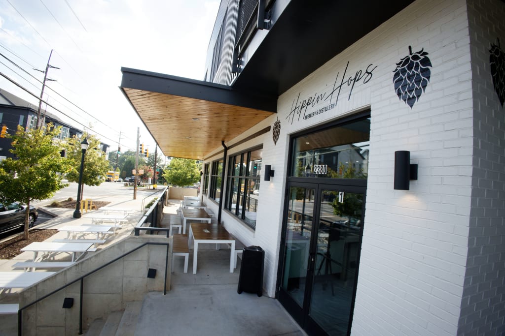 A modern brewery with a white brick facade and wooden roof overhang features outdoor seating and large windows in Atlanta, Georgia.