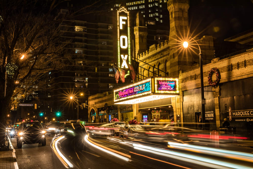 A vibrant city street at night, illuminated by car lights and a theater marquee in Atlanta, Georgia.