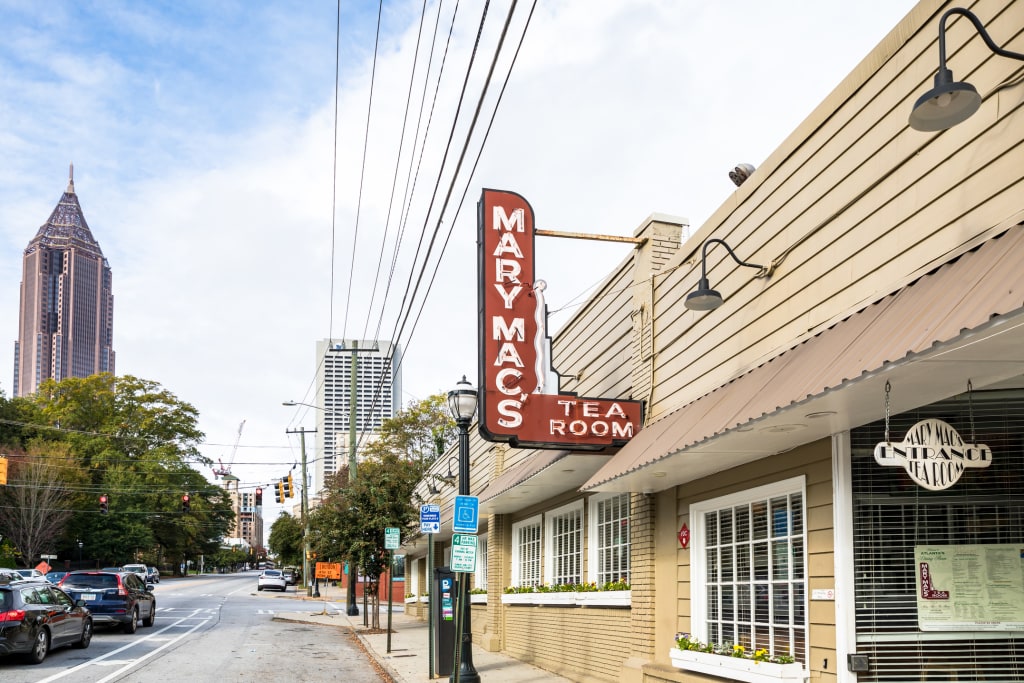 A quaint tea room with a vintage sign stands on a city street lined with trees and modern skyscrapers in Atlanta, Georgia.