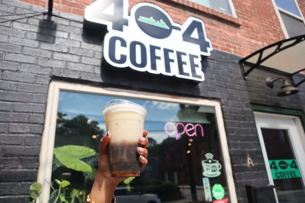 Hand holding a cold coffee drink outside a brick coffee shop with a neon open sign in Atlanta, Georgia.