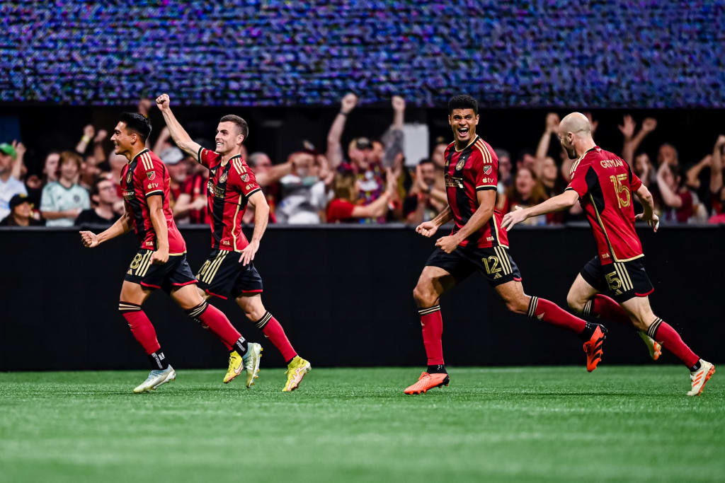 Four soccer players in red and black uniforms celebrate a goal with fans in the background in Atlanta, Georgia.