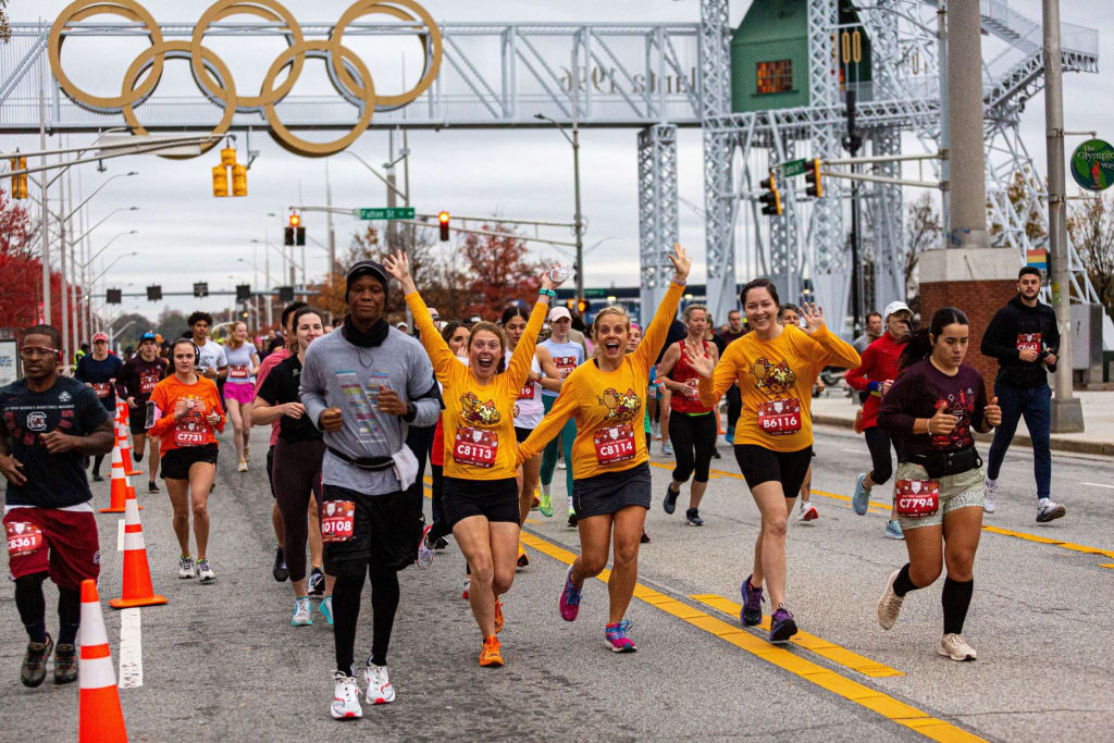 A large group of runners in colorful athletic wear participate in a marathon, passing under the Olympic rings in Atlanta, Georgia.