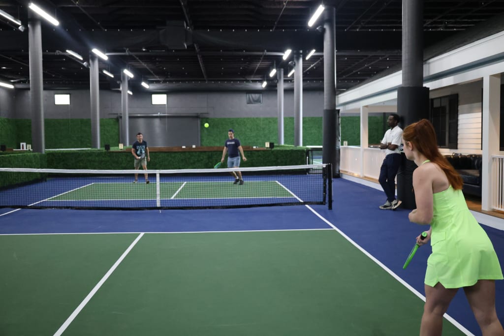 Four people play tennis on an indoor court with green walls and blue flooring in Atlanta, Georgia.
