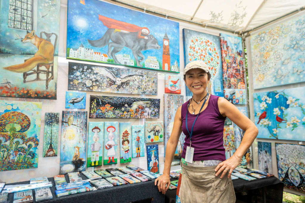 Artist stands in front of vibrant, whimsical paintings and prints at an outdoor art fair in Atlanta, Georgia.