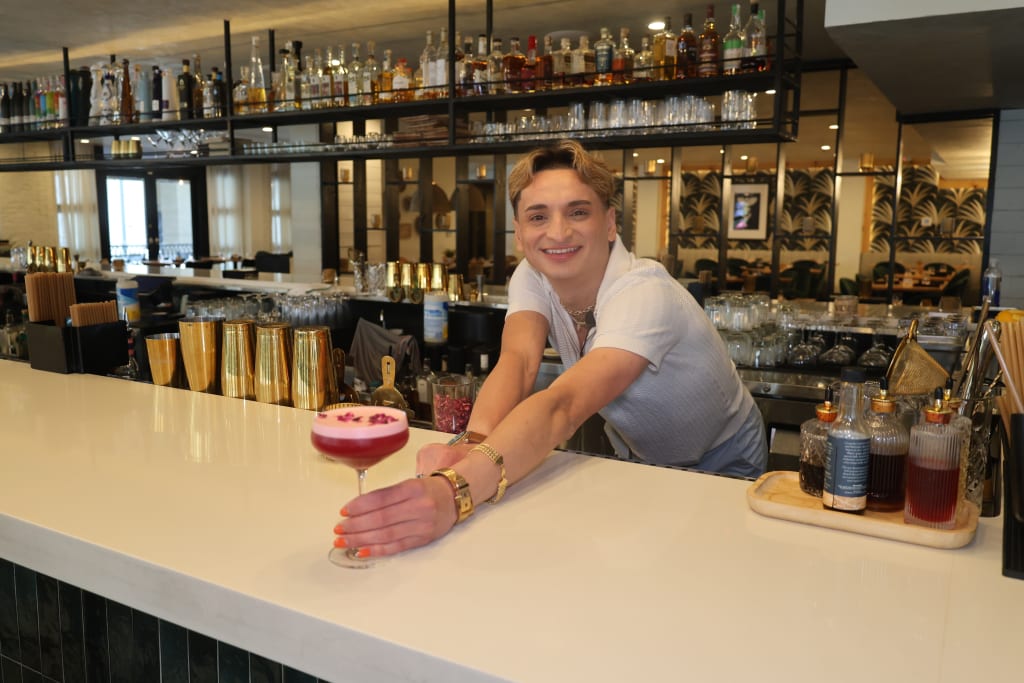 A smiling bartender presents a colorful cocktail at a modern bar with shelves of liquor in Atlanta, Georgia.
