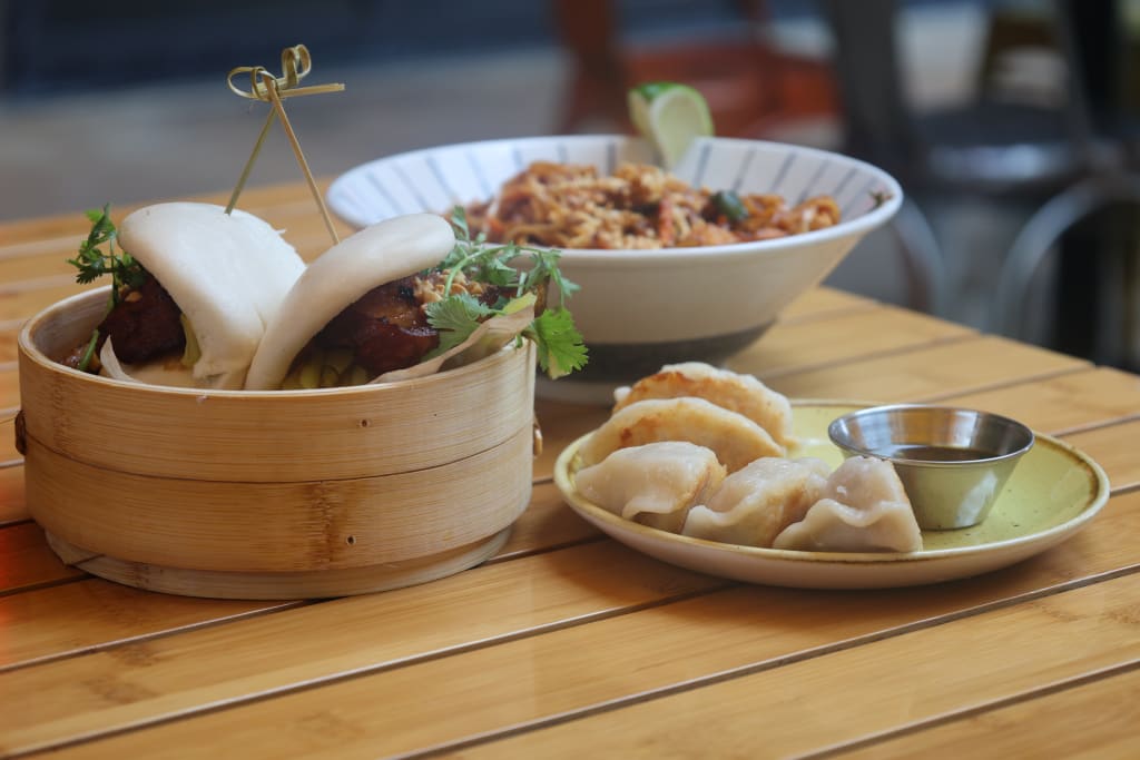 Explore Atlanta Beltline Eastside Trail 15 A wooden table holds a bamboo steamer with pork belly bao buns, a plate of fried dumplings, and a bowl of noodles in Atlanta, Georgia.