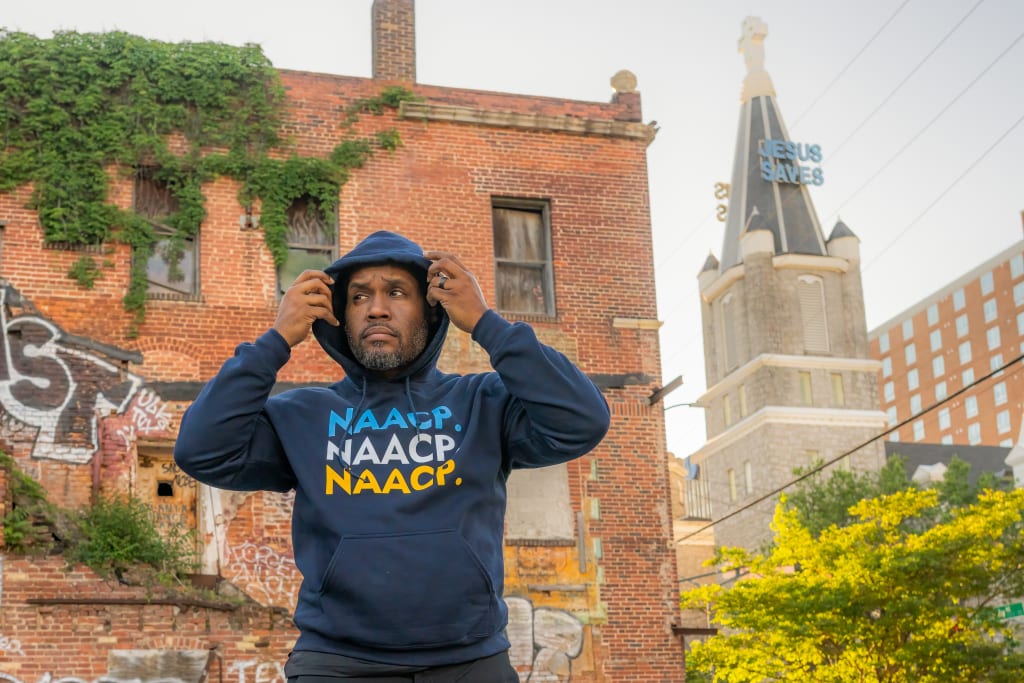 A man stands in front of a brick building with graffiti, wearing a hoodie with the letters NAAACP in Atlanta, Georgia.