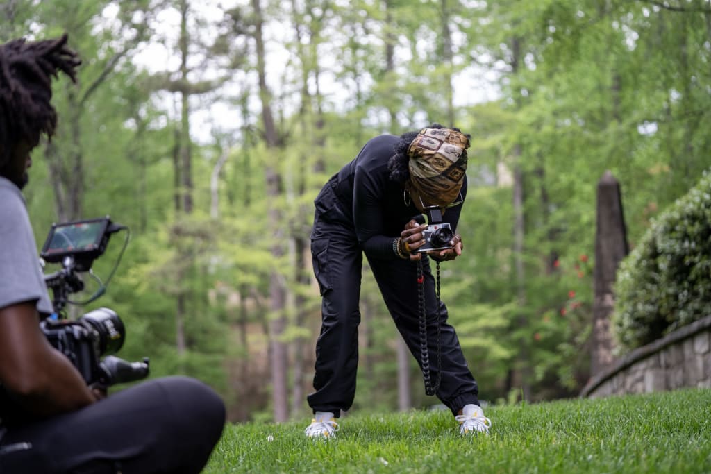 A person in black clothing and a bandana is crouching on grass, holding a camera in Atlanta, Georgia.