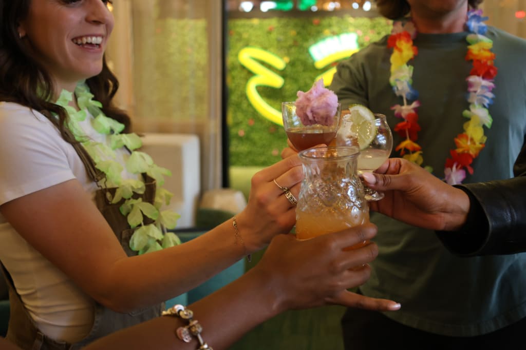 Three people cheerfully clink glasses filled with colorful drinks, wearing festive leis in Atlanta, Georgia.