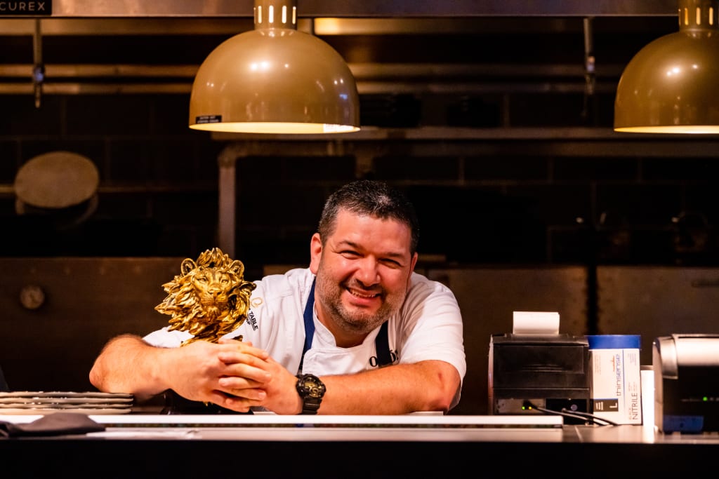 A smiling chef proudly displays a golden lion head trophy in a dimly lit kitchen in Atlanta, Georgia.