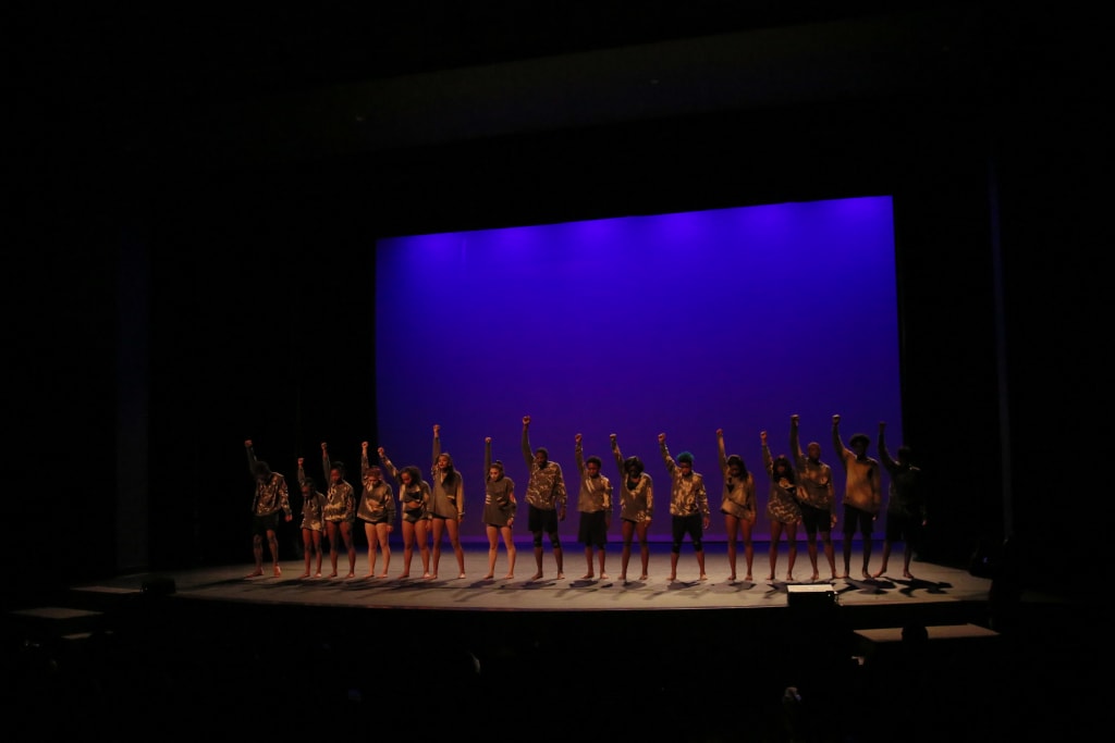 A group of dancers perform on stage, raising their arms in unison against a purple backdrop in Atlanta, Georgia.